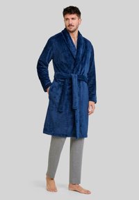 Bata de baño texturizada en azul marino con cuello chal, cinturón, bolsillos laterales, llevada sobre pantalones grises claros. Tejido suave con un patrón de cuadros.