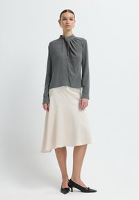 Graues Langarmoberteil mit einem geknoteten Detail am Kragen, kombiniert mit einem fließenden elfenbeinfarbenen Rock und schwarzen spitzen High Heels.