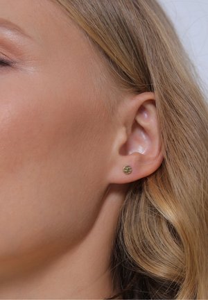 Oreille gauche d'une femme avec une petite boucle d'oreille ronde en or texturé et cheveux blonds repliés derrière l'oreille sur un fond clair.