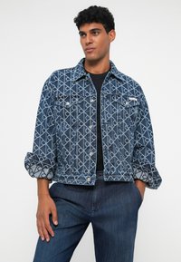Moški nosi modro vzorčasto denim jakno s zavihki čez črno srajco in modre kavbojke, stoji z eno roko v žepu.