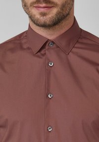 Bruine button-up blouse met een gladde textuur, voorzien van een puntige kraag en vier metalen knopen met rode accenten.