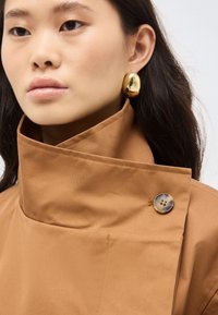 Femme aux longs cheveux bruns portant des boucles d'oreilles épaisses en or et un manteau marron à col haut fermé par un gros bouton près du cou.