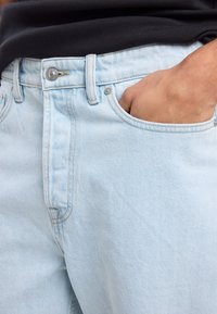 Vaaleansiniset farkut suoralla punoksella, joissa on etupainike, päällä ommellut taskut ja pehmeä, teksturoitu pinta.