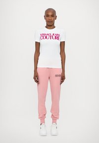 Valge kitsas T-särk, millel on roosa kiri "VERSACE JEANS COUTURE", koos heleda roosa dressipluusiga, millel on sama logo. Valged tossud.