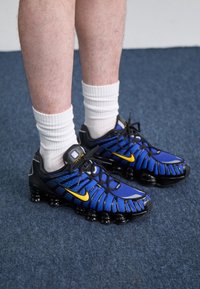 Modré a černé sportovní boty s texturovaným vrškem, žlutými akcenty a síťovanými detaily, spárované s bílými žebrovanými ponožkami.