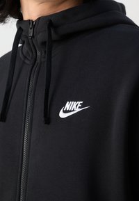 Schwarze Zip-Up-Hoodie aus weichem Stoff, mit hohem Kragen, Kordeln und weißem Nike-Logo auf der Brust. Glatte Textur.