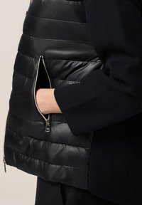Sort puffer vest med en skinnende tekstur, der har en sidelomme med lynlås og vandret quiltet design, parret med en mørk strikket ærme.