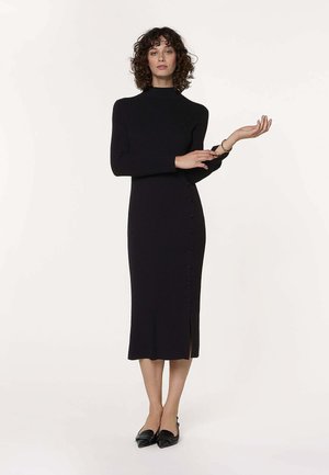 Femme aux cheveux bouclés portant une robe midi noire à manches longues avec des boutons sur le côté et des ballerines noires pointues, posant avec un bras levé.