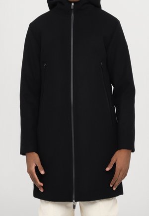 Osoba ubrana w czarny płaszcz z kapturem z przednim zamkiem błyskawicznym i zapinanymi bocznymi kieszeniami, w jasnych spodniach, stojąca na jednolitym tle.