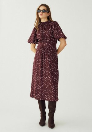 Kobieta w czarnych okularach przeciwsłonecznych, bordowej sukience midi z bufkami i kwiatowym printem oraz ciemnobrązowych kozakach do kolan, stoi z rękami za plecami.