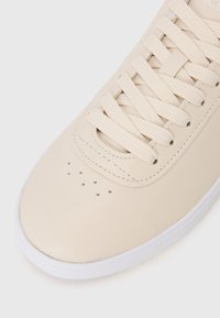 Beige lærersneaker med flate lisser, perforert tåboks og hvit gummisåle. Glatt overflate og minimalistiske designdetaljer.