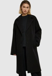 Svart ullkåpe med bred krage, to knapper, sidelommer og en oversized passform. Tekstruert stoff og minimalistisk design.