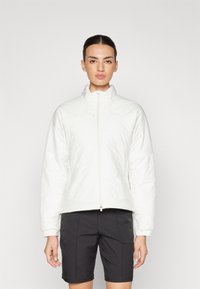 Balta paminkštinta striukė su aukštu apykakle, užtrauktuku ir ilgomis rankovėmis. Lygus paviršius su šiek tiek blizgiu apdailos efektu. Suderinta su tamsiomis šortais.