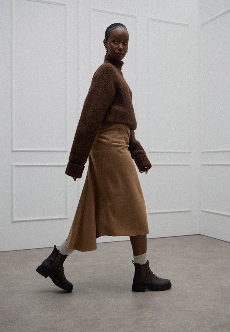 Suéter de punto marrón, falda midi color camel, botas negras de tobillo con suela texturizada, llevadas con calcetines blancos. Fondo de habitación minimalista.
