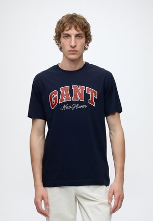 Junger Mann mit lockigem Haar trägt ein dunkelblaues GANT New Haven T-Shirt und helle Hosen und steht vor einem schlichten Hintergrund.