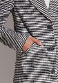 Zwart-wit houndstooth patroon jas met een notch-lapel, grote zwarte knopen en zijzakken. Textuurafwerking van de stof.