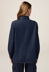 Camicia in denim scuro con colletto a punta, maniche lunghe e polsini con bottone. Presenta uno yoke sul retro e dettagli di cucitura sottili.