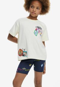 Camiseta de manga corta crema con gráficos de parches coloridos y acentos bordados. Pantalones cortos oscuros con patrones vibrantes y juguetones.