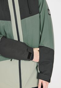 Groene en zwarte jas met een hoge kraag, een rits aan de voorkant en verstelbare manchetten. Gemaakt van gestructureerd, weerbestendig materiaal met logo-details.