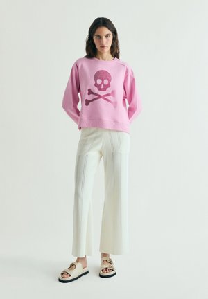 Mujer de pie, vestida con un suéter rosa con un gráfico de calavera y huesos cruzados, pantalones blancos de pierna ancha y sandalias beige.