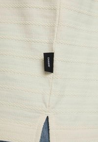 Hellbeige Pullover mit strukturierten Streifen und seitlichem Schlitz, ausgestattet mit einem schwarzen Etikett mit weißem Text. Weiches Strickmaterial.