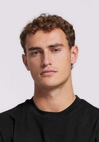 Zwarte t-shirt met een ronde hals, gemaakt van een zachte, gestructureerde stof. Model heeft kort, krullend haar en een neutrale huidtint.