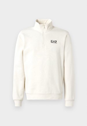 Krém színű, negyed cipzáras pulóver hosszú ujjakkal, bal mellen EA7 Emporio Armani logóval, egy sima háttér előtt.