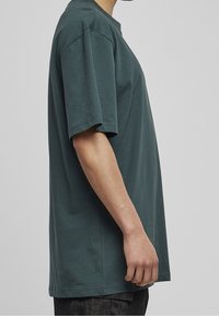 Persona in una maglietta oversize a maniche corte verde scuro e pantaloni neri, mostrata di profilo su uno sfondo grigio chiaro semplice.