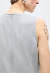 Άτομο που φοράει αμάνικο γκρι ανοιχτό τοπ με φερμουάρ στην πλάτη, δείχνοντας κοντά μαλλιά, αφηρημένο τατουάζ στον άνω βραχίονα και μεγάλο διαφανές σκουλαρίκι.