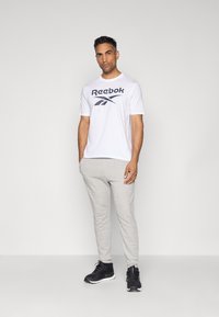 Weißes T-Shirt mit navyblauem Reebok-Logo, kombiniert mit hellgrauen Jogginghosen und schwarzen Sneakers. Einfaches Design, lockere Passform, glatte Textur.