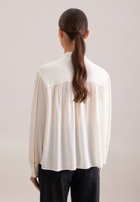 Blouse blanche fluide avec un col boutonné, des manches longues et des détails plissés dans le dos, associée à un jean en denim foncé.