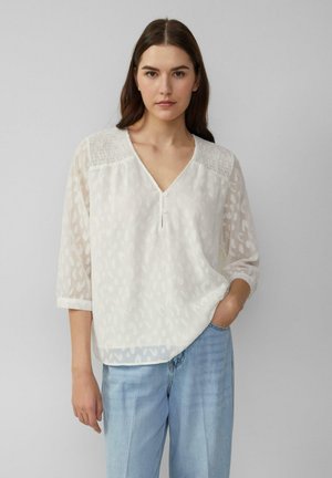 Blouse semi-transparente blanche avec un col en V, des manches trois-quarts et un motif ton sur ton subtil. Présente une texture froncée au niveau des épaules.