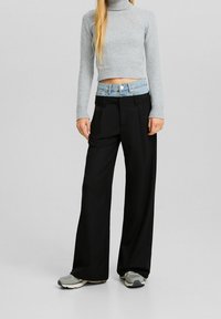 Person som bär lager av svarta vida byxor över blå jeans, grå kortärmad tröja med polokrage och grå sneakers mot en neutral bakgrund.