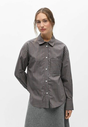 Femme portant une chemise à carreaux marron et grise boutonnée et une jupe grise, debout devant un fond blanc uni.