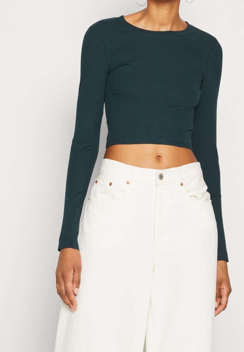 Tiefgrünes, geripptes Langarm-Crop-Top mit rundem Ausschnitt, kombiniert mit hoch taillierten, weißen weiten Hosen mit Kupferknopf-Akzenten.