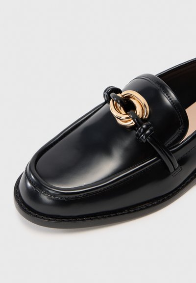 Black leather loafer with a glossy finish, featuring a gold interlocking ring detail, rounded toe, and decorative stitching along the edges.