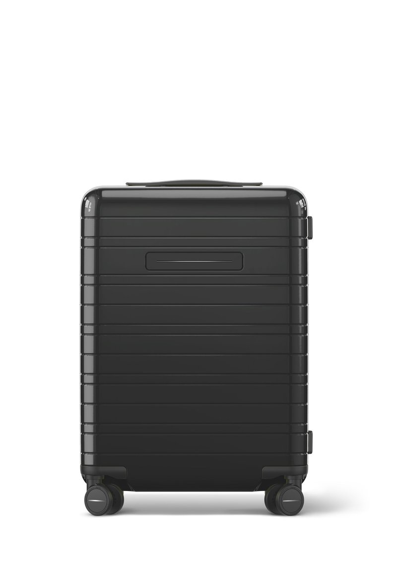 Valise rigide noire avec des rainures horizontales, une poignée supérieure et quatre roulettes pivotantes pour la mobilité. Texture lisse et forme rectangulaire.