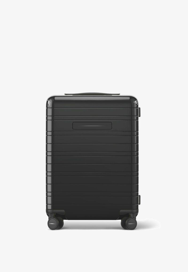 Valise rigide noire avec des rainures horizontales, une poignée supérieure et quatre roulettes pivotantes pour la mobilité. Texture lisse et forme rectangulaire.