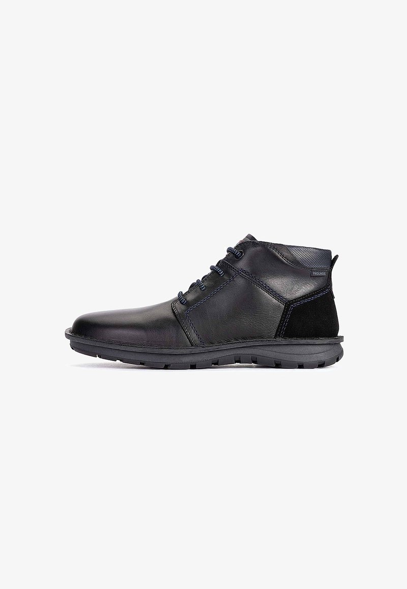 Bottine en cuir noir avec laçage à l'avant, col matelassé, semelle texturée et détails cousus sur fond blanc.
