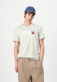 Light green cotton t-shirt with "TOMMY" logo in white, red, and navy. Paired with beige pleated trousers and a navy cap.