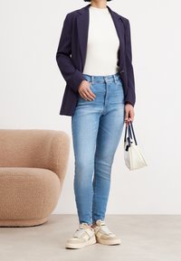 Blazer azul marino, top blanco de canalé, jeans ajustados azul claro, zapatillas beige con correas, sosteniendo un pequeño bolso blanco con acentos azul marino.