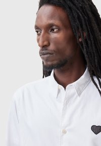 Camisa blanca de botones hecha de algodón con un corazón negro texturizado bordado en el lado izquierdo del pecho. Cuenta con un cuello estándar y mangas largas.