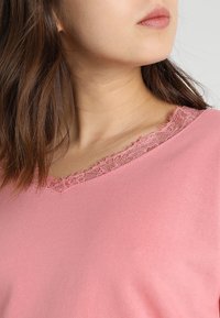 Růžový top s výstřihem do V a texturovaným krajkovým lemem kolem výstřihu; měkká tkanina s hladkým povrchem, přiléhavý střih a krátké rukávy.