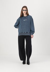 Pulover iz denima v temno modri barvi z rebrastimi manšetami in robom, ki vsebuje bel odtis logotipa. V kombinaciji z širokokrajnimi črnimi hlačami in črnimi gležnjarji.