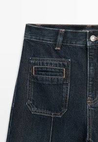Dark blue denim jeans with a classic design, featuring a front pocket, a rectangular back pocket with stitching detail, and a metal button closure.