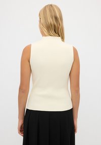 Mouwloze ribbed top in crème met een hoge hals, aansluitende silhouet en een gladde textuur. G gedragen met een zwarte geplooide rok, minimalistisch ontwerp.