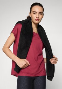 Top rosso a maniche corte abbinato a un outer layer senza maniche di colore nero e texture. L'outer layer ha una finitura morbida e pelosa. Leggings scuri a fantasia completano l'outfit.