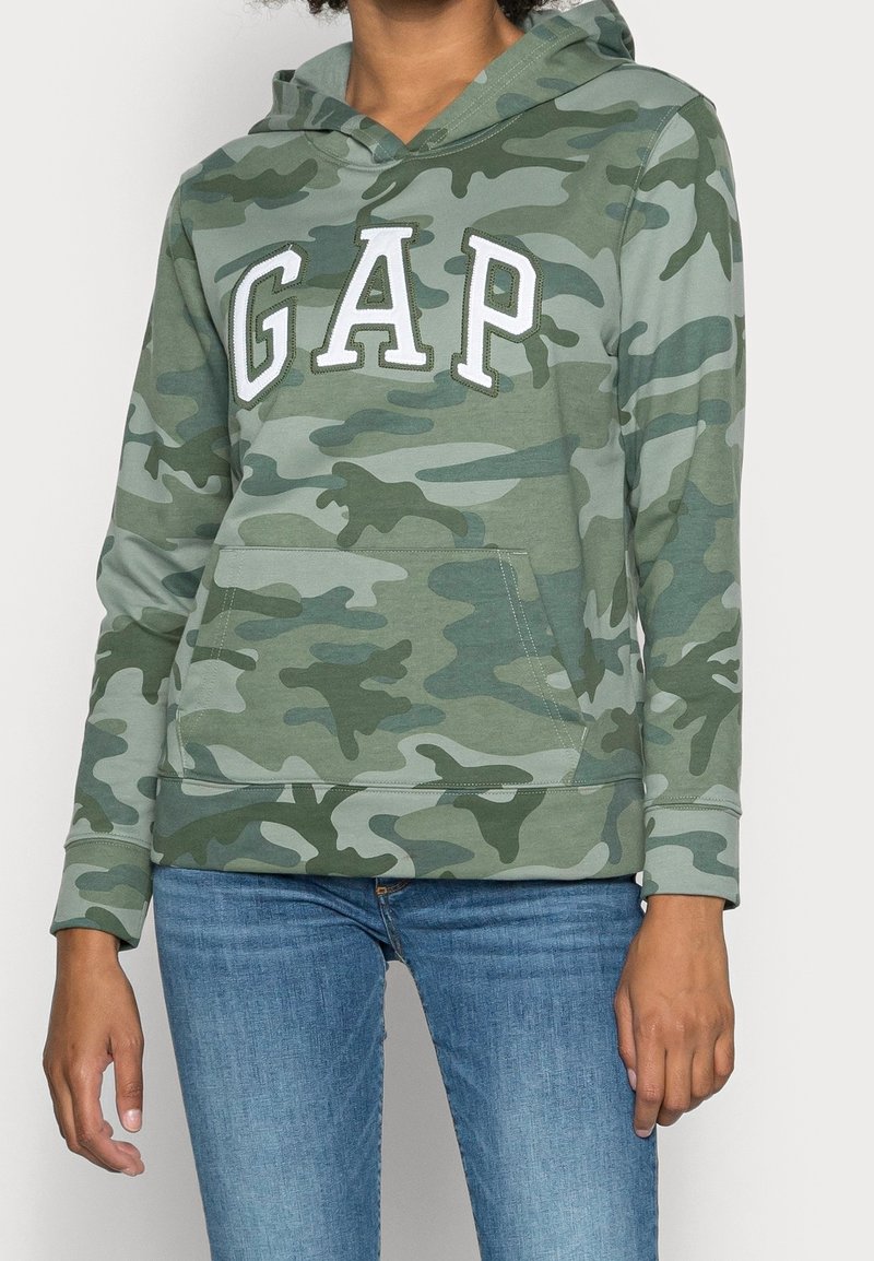 Personne portant un sweat à capuche camouflage vert avec les lettres blanches "GAP" et un jean bleu, visible des épaules aux hanches.