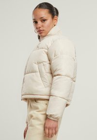 Beige puffjakke med en kort design, polstret tekstur og elastiske mansjetter. Har sidelommer og høy krage.