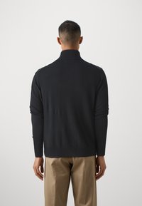 Suéter preto de malha com gola alta, design ajustado e mangas longas, combinado com calças bege, visto de costas.